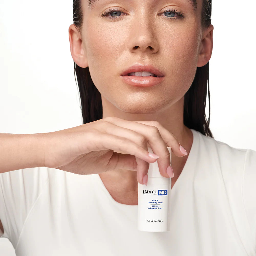 Eine Frau mit minimalem Make-up und nassem Haar hält eine kleine weiße Flasche Image Skincare - IMAGE MD gentle cleansing balm vor ihrer Brust, vor einem schlichten weißen Hintergrund.