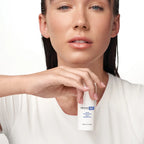 Eine Frau mit minimalem Make-up und nassem Haar hält eine kleine weiße Flasche Image Skincare - IMAGE MD gentle cleansing balm vor ihrer Brust, vor einem schlichten weißen Hintergrund.