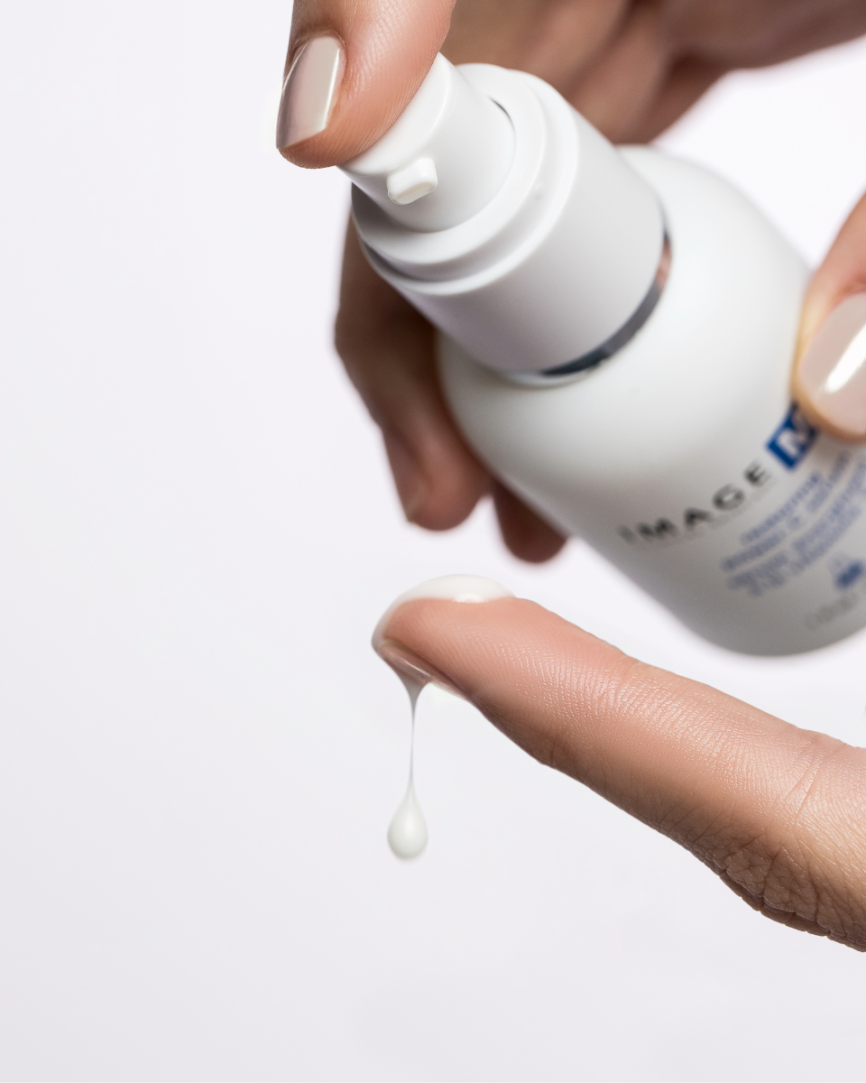 Eine Hand spendet das IMAGE MD Restoring Power C Serum von Image Skincare aus der Flasche auf eine Fingerspitze, wobei sich zwischen Spender und Finger ein weißer Cremetropfen vor einem schlichten weißen Hintergrund bildet.