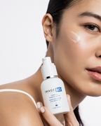 Eine Frau mit strahlender Haut trägt das IMAGE SKINCARE IMAGE MD Restoring Power C Serum auf ihre Wange auf. Sie hält die weiße Flasche mit der Aufschrift Image Skincare und trägt ein weißes Armband.