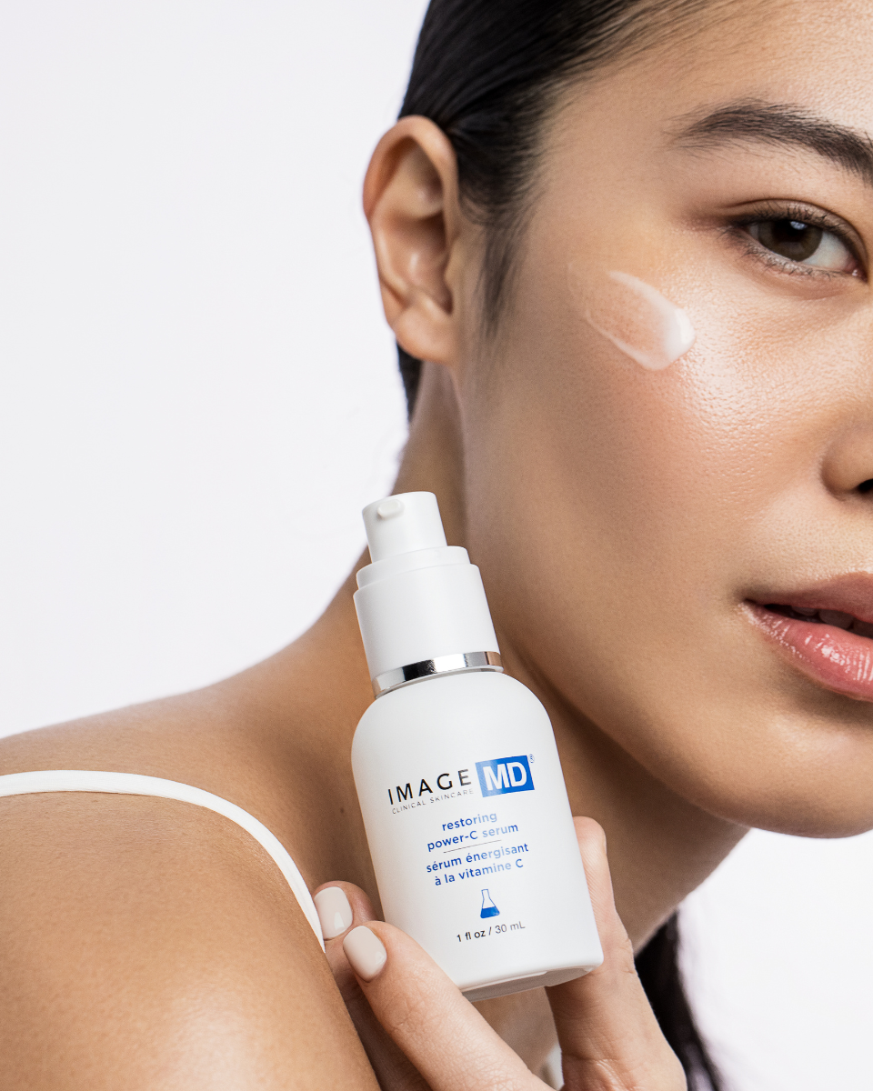 Eine Frau mit strahlender Haut trägt das IMAGE SKINCARE IMAGE MD Restoring Power C Serum auf ihre Wange auf. Sie hält die weiße Flasche mit der Aufschrift Image Skincare und trägt ein weißes Armband.