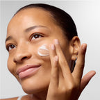 Eine Frau mit reiner Haut lächelt sanft, während sie vor einem schlichten Hintergrund mit den Fingern eine kleine Menge des „dermalogica – Stabilizing Repair Serum“ auf ihre Wange aufträgt, um zu verdeutlichen, wie wichtig die Verwendung des dermalogica-Serums für empfindliche Haut zur Stärkung der Hautbarriere ist.