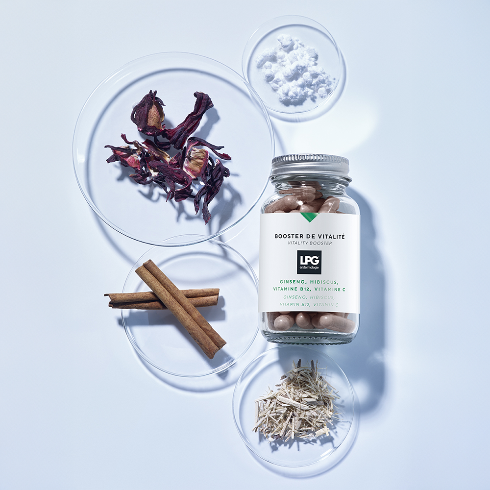 Ein Glas mit LPG - Vitality Booster Nahrungsergänzungskapseln von LPG steht auf einem hellblauen Hintergrund, umgeben von Glasschalen mit getrocknetem Hibiskus, Zimtstangen, weißem Pulver und zerkleinerten Wurzeln.