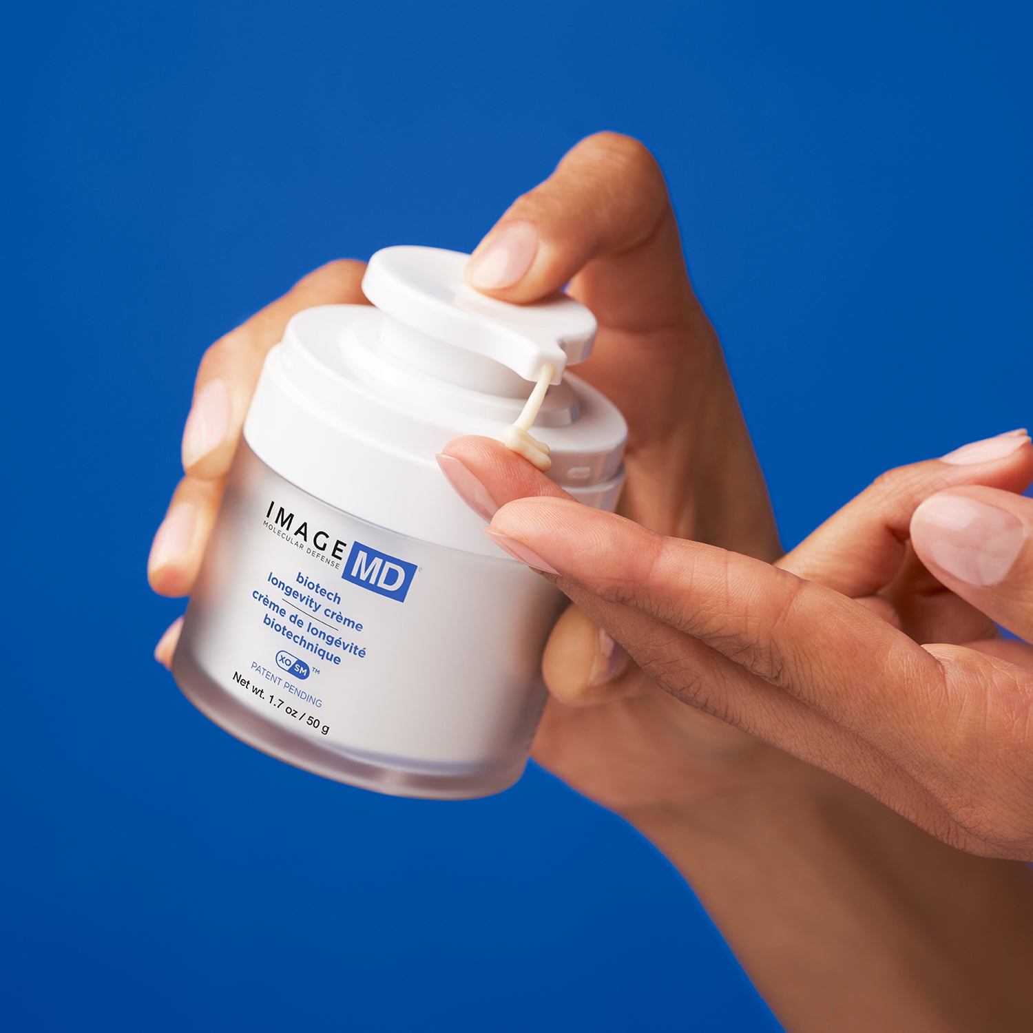 Eine Person gibt eine kleine Menge der Image Skincare - IMAGE MD biotech longevity crème auf ihre Fingerspitze aus einer weißen Pumpflasche vor einem einfarbig blauen Hintergrund ab.