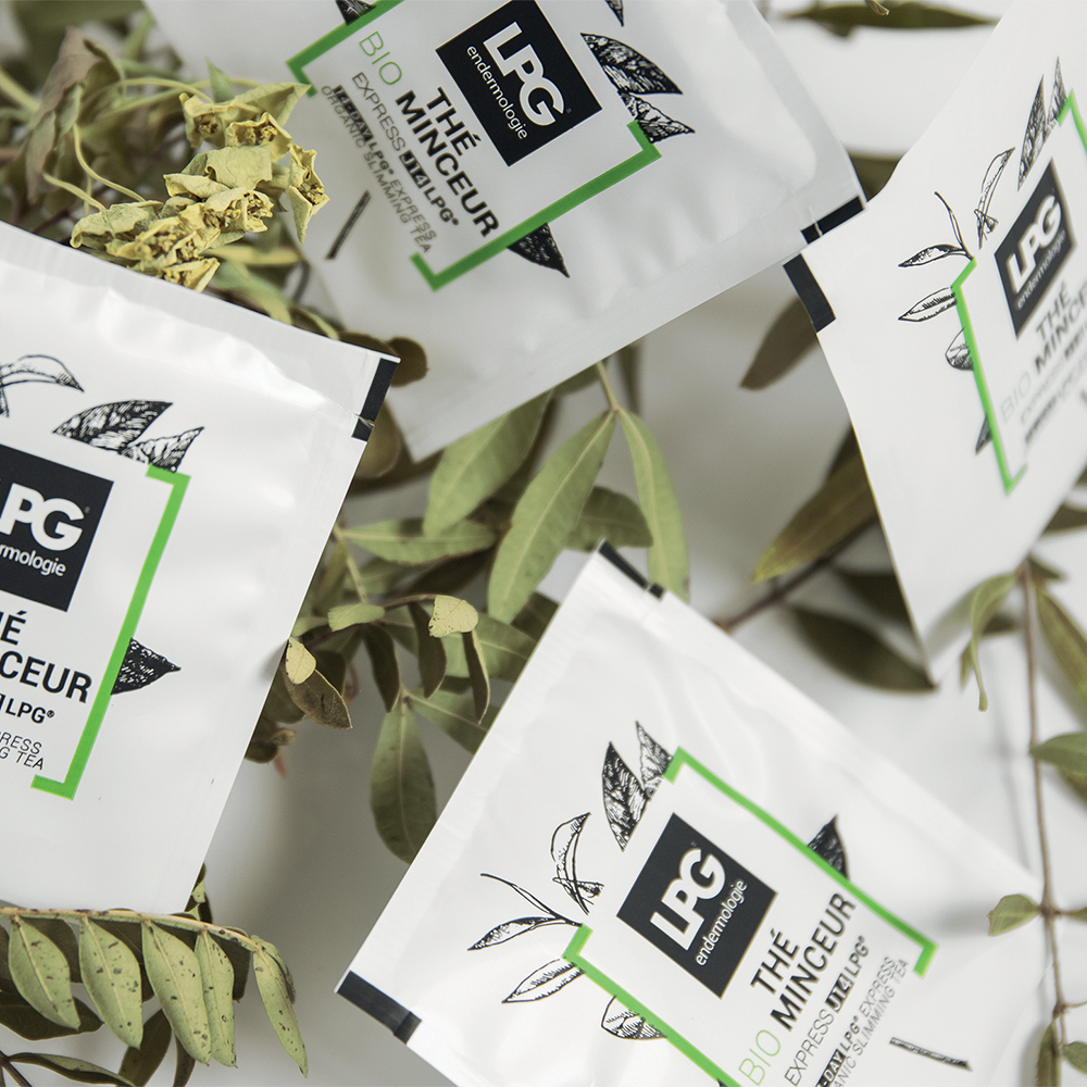 Mehrere Tütchen LPG - 14 Tage Express Bio Abnehmtee sind auf belaubten Zweigen ausgestellt. Die weißen Tütchen mit botanischen Illustrationen und grünen Akzenten tragen auf der Vorderseite den Markennamen LPG und Angaben zum Produkt.